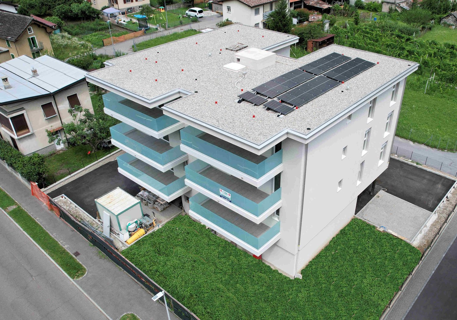 A modern apartment building with four floors and glass balconies. The building is made of white and blue materials, with solar panels on the rooftop. The building has multiple windows and is surrounded by grass, trees, and a road. In the distance, there a