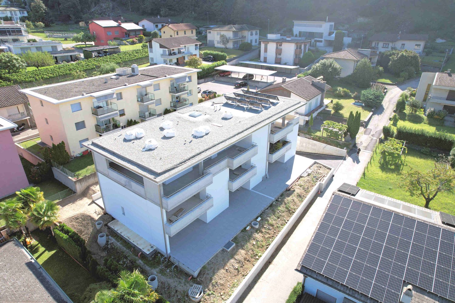 4-story residential building, modern architecture, balconies, solar panels on the roof, covered parking area, communal garden, surrounding houses and greenery