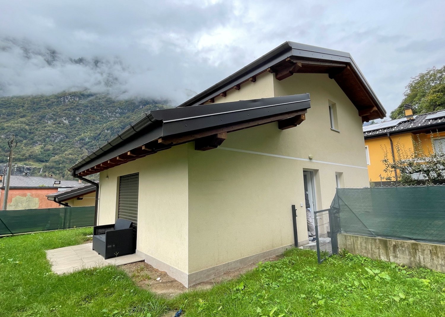 single-story house, beige exterior, black roof, outdoor seating area, surrounded by green grass, mountains in the background