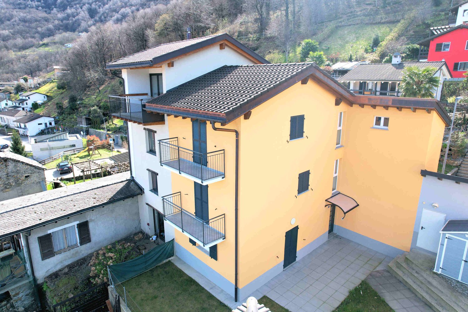 Two-story building with balconies, yellow exterior, tiled roof, multiple windows, and entrance with awning, surrounded by other buildings and trees