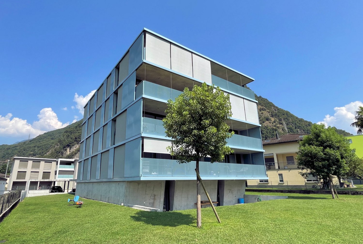 multi-storey building, blue exterior, multiple balconies, located in front of a mountain, fenced area, green lawn