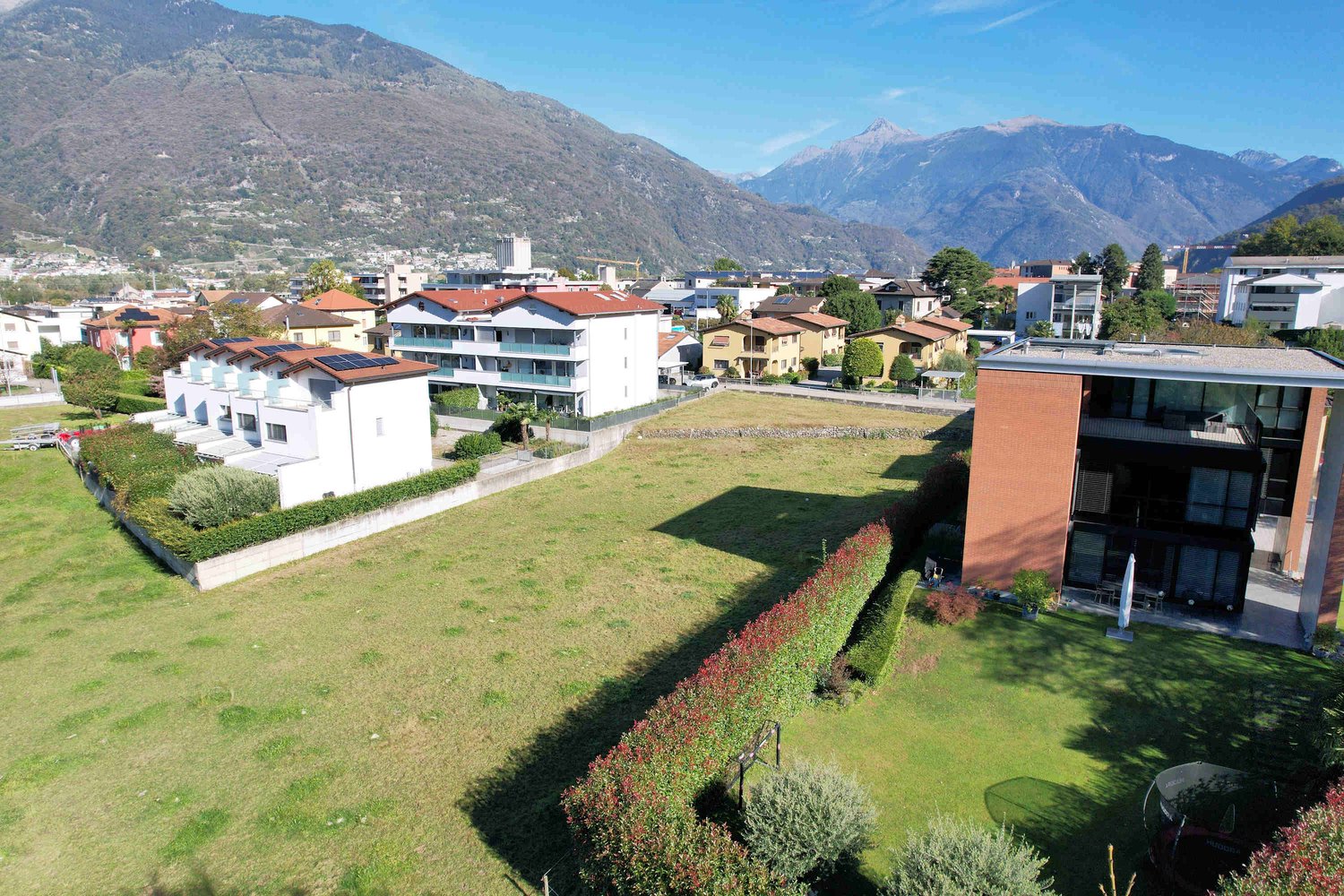 Apartment buildings with red roofs, large open grassy area, surrounding greenery, mountain backdrop