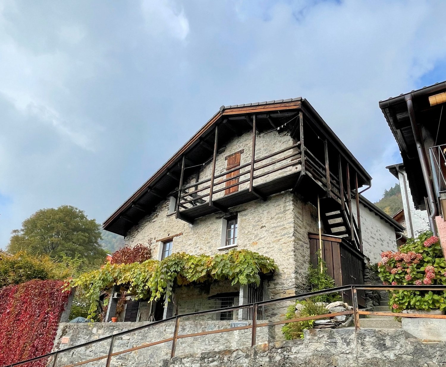 stone wall, wooden balcony, wooden door, wooden staircase, plants on the wall, red leaves, mountain in the background