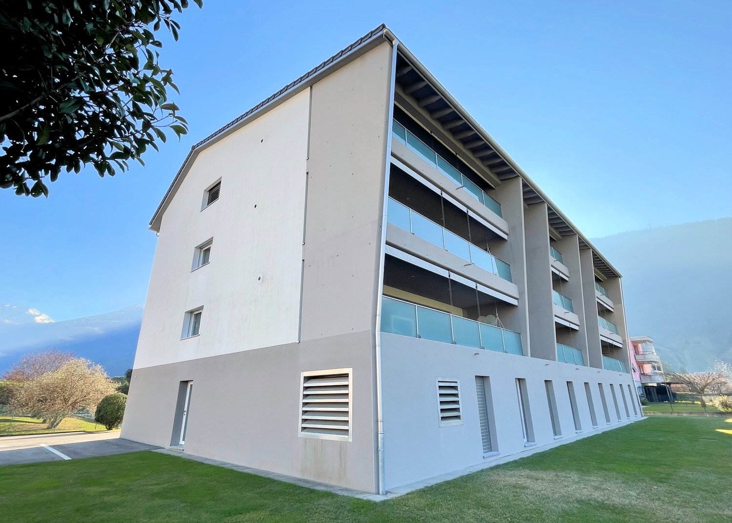 Modern apartment building with multiple floors, glass windows, balconies, and a green lawn in front.