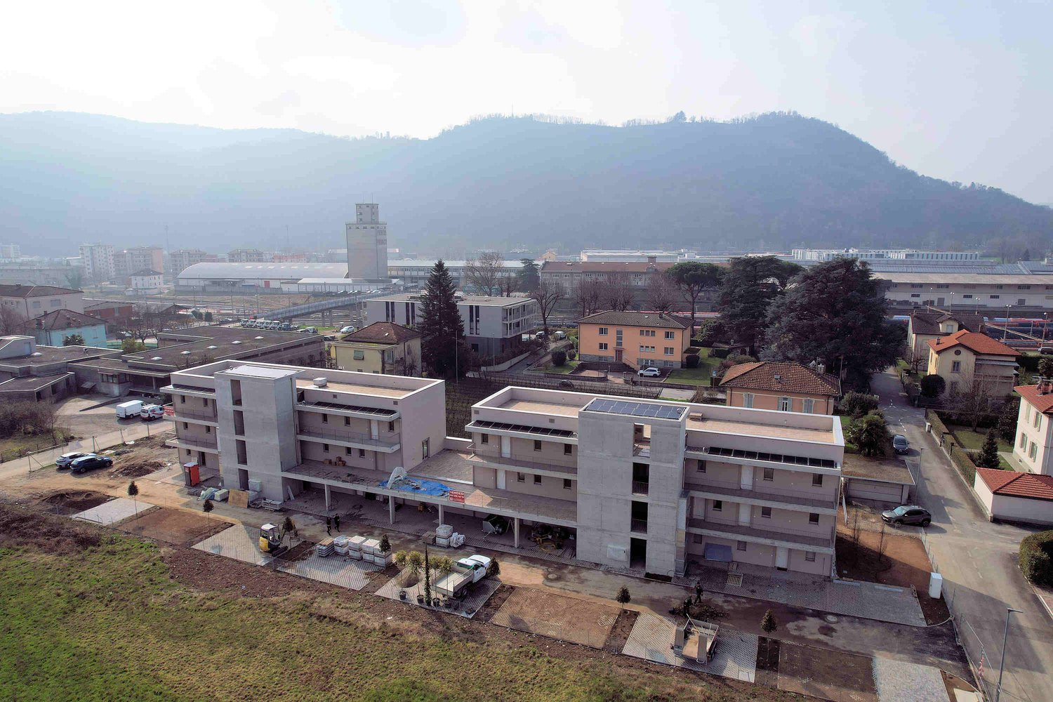 Aerial view of a multi-story building under construction, surrounded by other buildings, trees, and a mountain in the background.