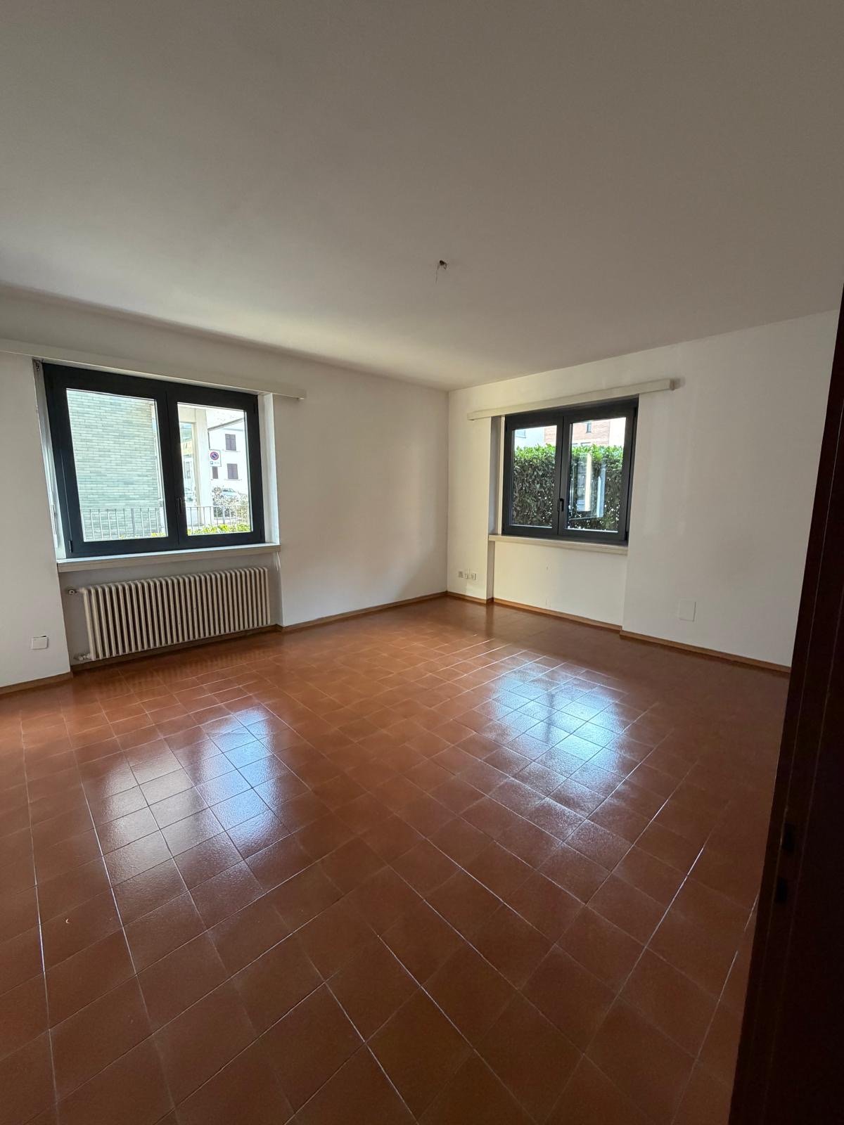 Empty room with tile flooring, white walls, two large windows with black frames, white radiators, and an electrical outlet.