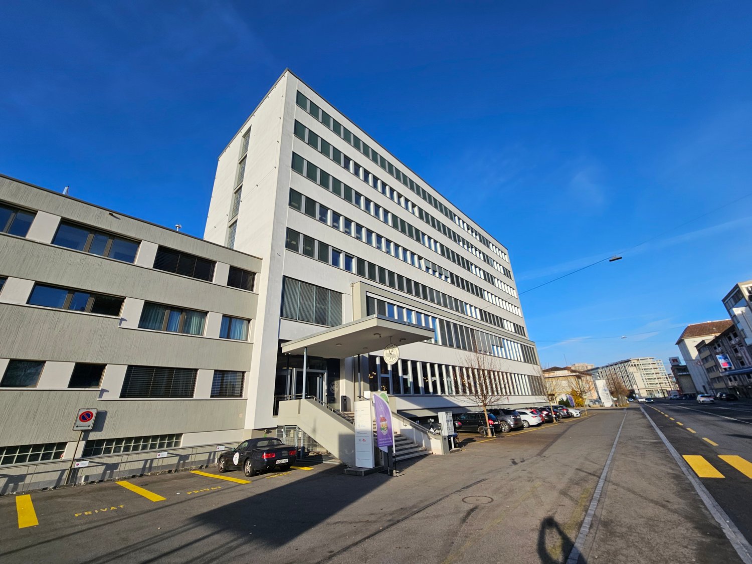 office building, multiple windows, 7 floors, car parked in front, stairs leading to entrance