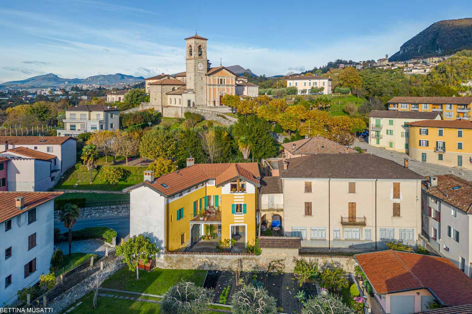 Aerial view of multiple buildings with varying architectural styles and colors set in a hilly landscape with a church tower visible in the background.