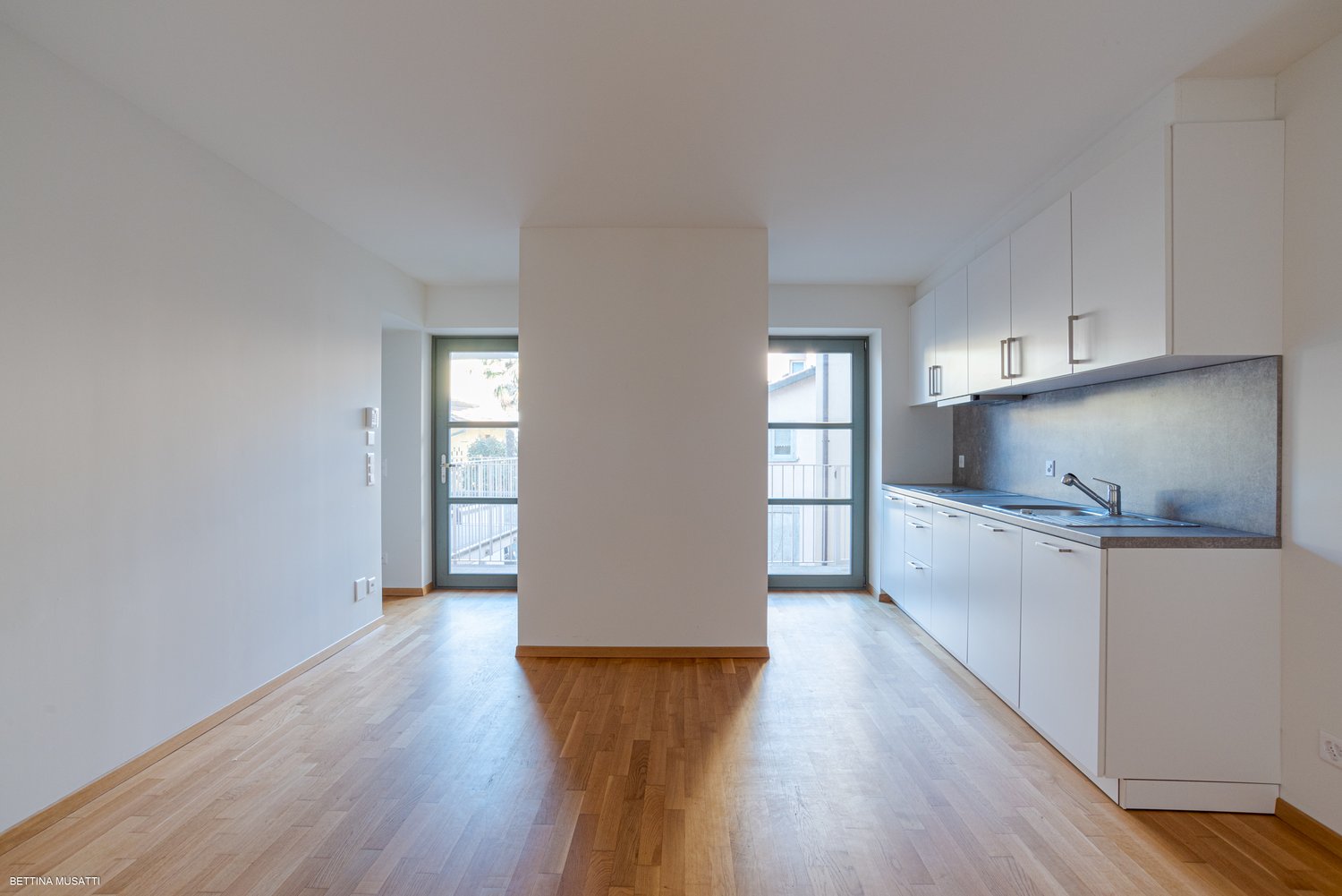 modern kitchen, wood floors, white walls, two large windows, central pillar, countertops, modern sink, faucet, built-in appliances