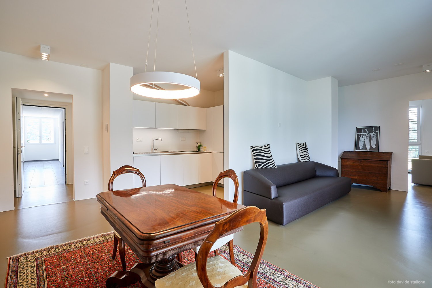 Open-concept living space with a dining area, kitchen, and living room. The dining table is made of wood with ornate chairs. The kitchen has white cabinets and a modern appliance, likely a dishwasher. The living room has a gray sofa and a decorative wall