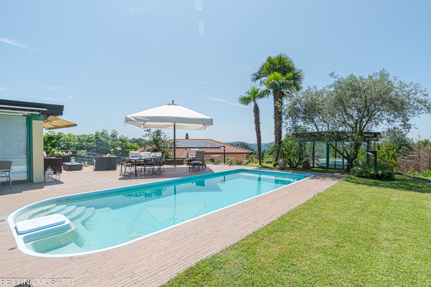 swimming pool, deck, lounge chairs, table, umbrella, grassy area, solar panels, trees