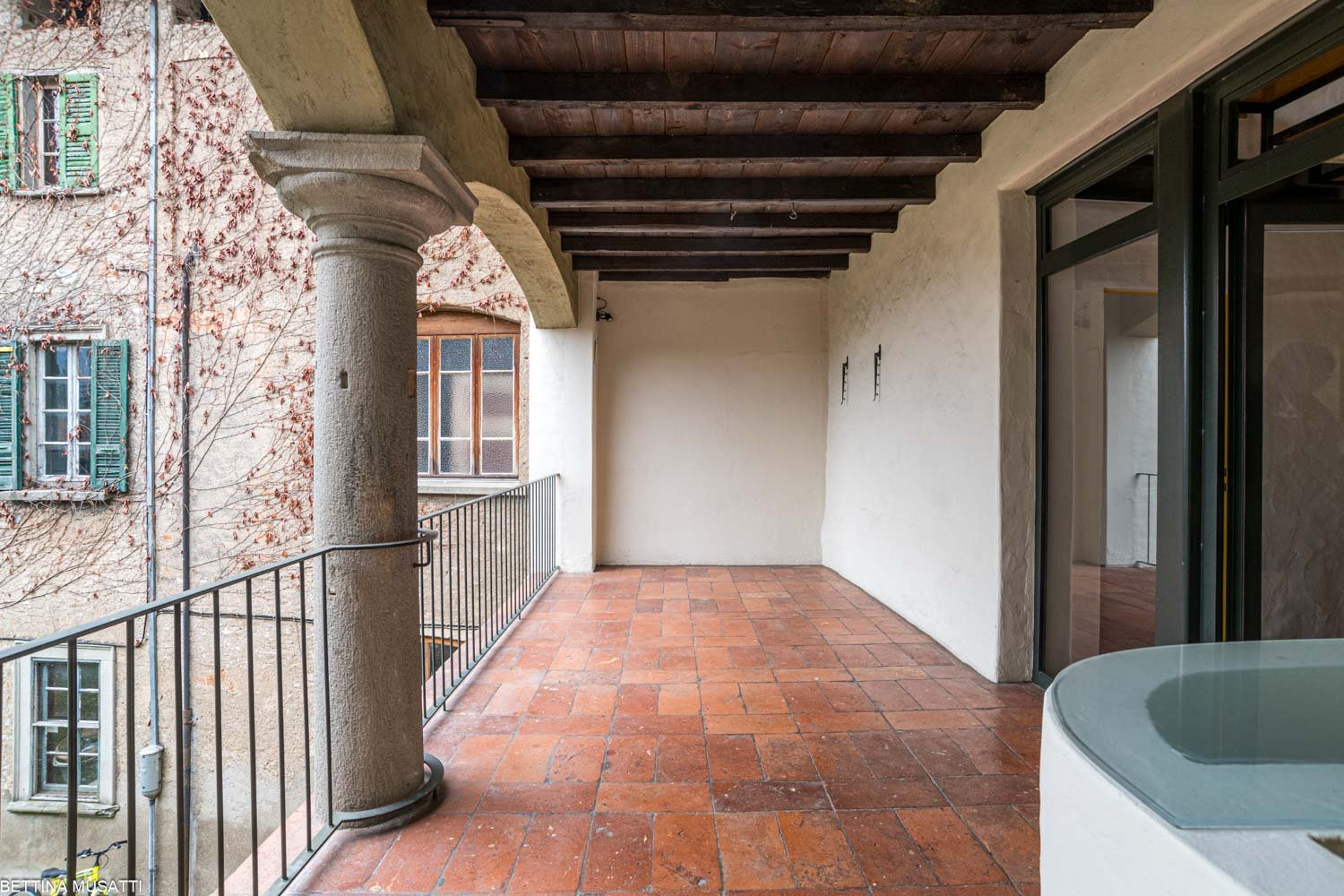 Covered balcony with stone columns, wooden beams, and terracotta tile flooring. The balcony overlooks a building with green shuttered windows.