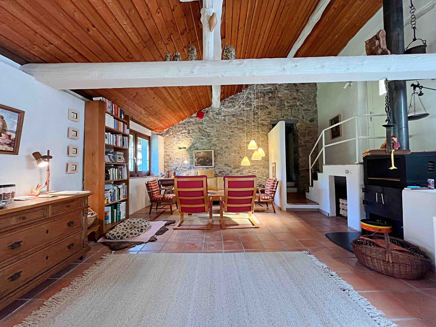 Rustic living room with exposed stone walls, wooden ceiling beams, a fireplace, bookshelves, a dining table with red chairs, and various decor items.