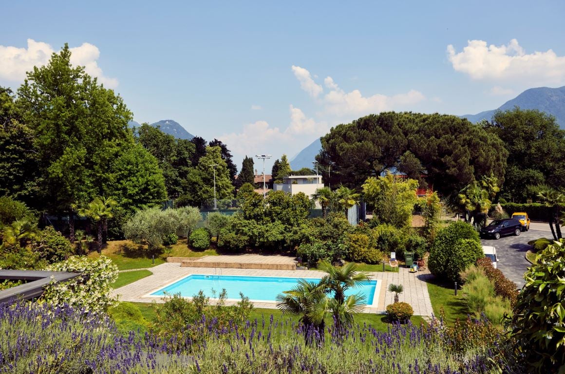 The image shows a large, well-maintained property with a swimming pool surrounded by lush greenery, including trees, shrubs, and a lavender garden. The property appears to have a balcony or terrace overlooking the pool and landscaped gardens. The backgrou