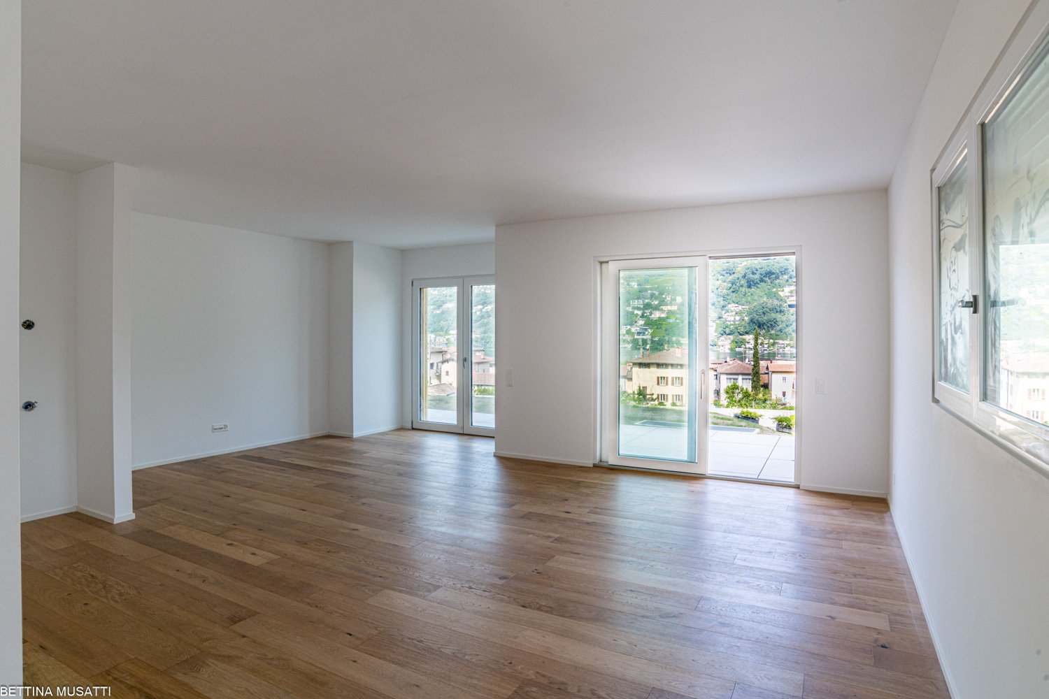 Spacious living room with large windows, hardwood flooring, and access to a balcony or terrace overlooking the surrounding neighborhood