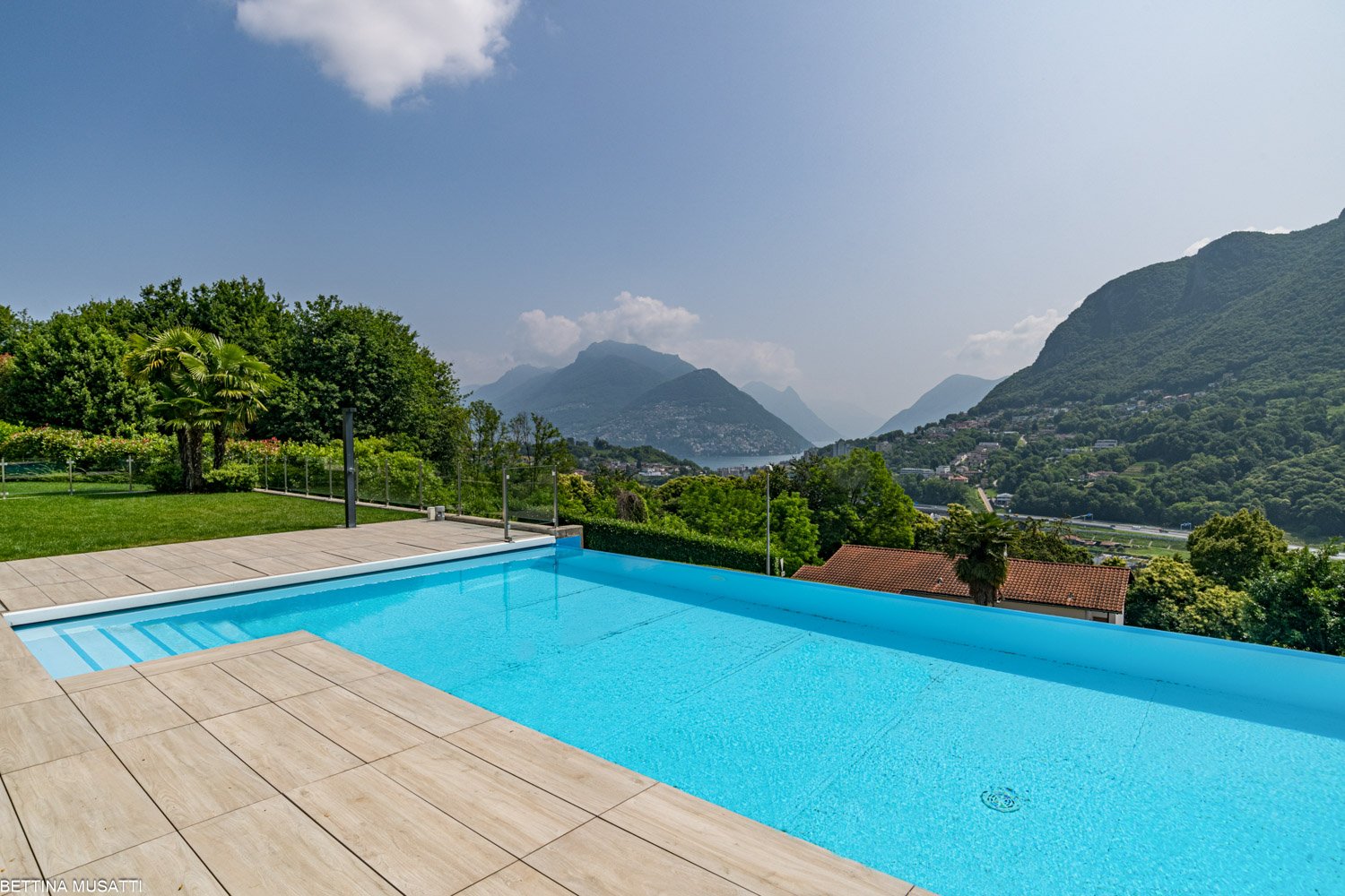Outdoor swimming pool with wooden deck, surrounded by lush greenery and with a scenic view of mountains and hills in the background.