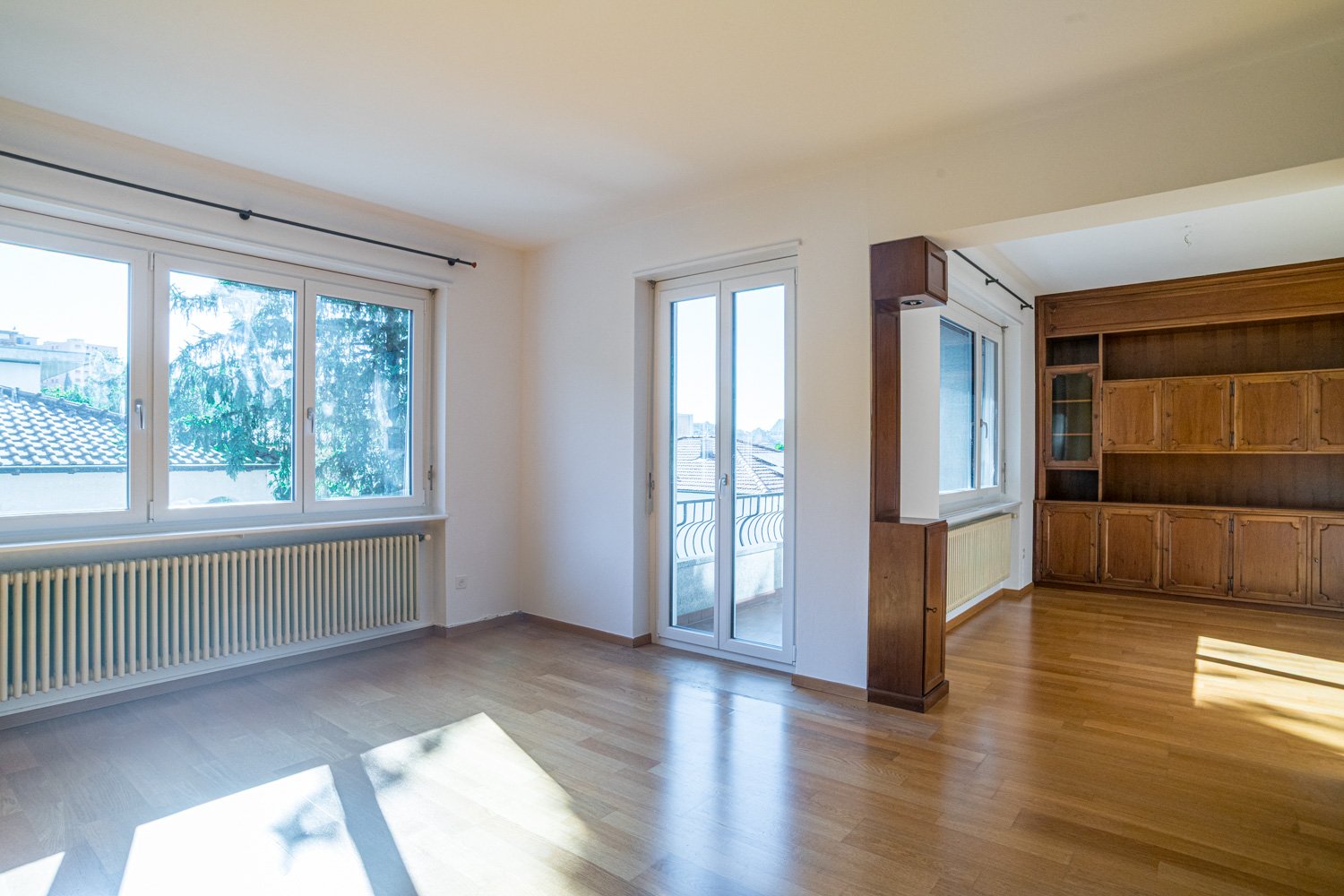 Spacious living room with large windows, hardwood floors, and a balcony or terrace overlooking the outdoor scenery. The room also features a built-in wooden cabinet or shelving unit.