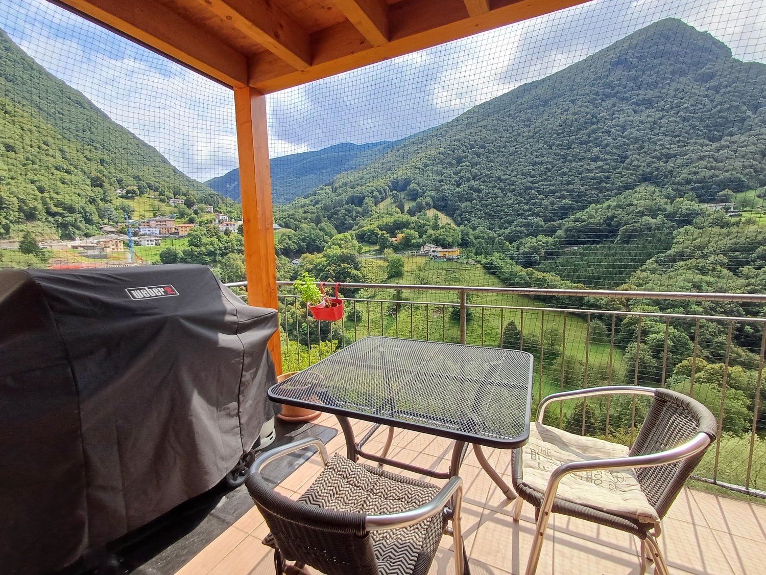 Furnished balcony with chairs, table, grill, plant, tiled floor, mesh railings, view of mountains and valley, hanging potted plant.