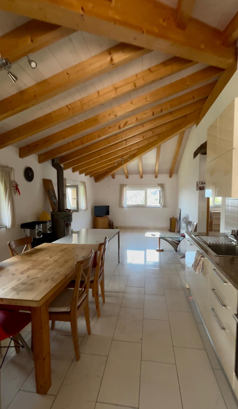 open dining area with wooden dining table, chairs, wooden beams on the ceiling, tiled floor, open kitchen with sink, appliances, small window with curtains, wooden cabinet, table and chair, black object, clock, and speaker.