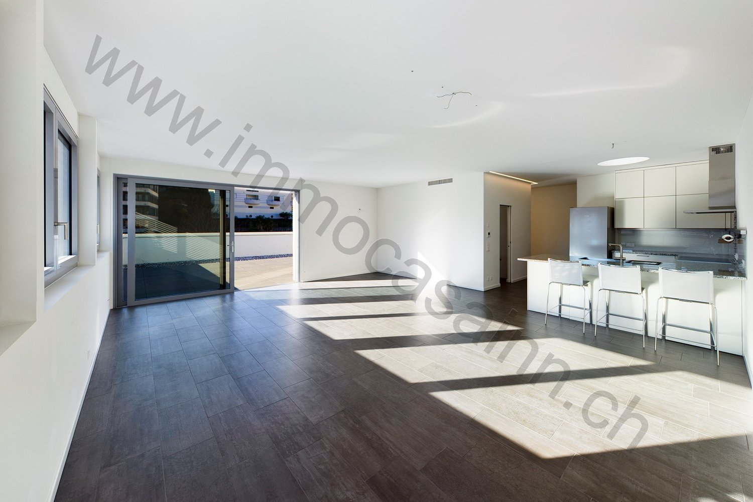 Spacious open-plan living area with hardwood flooring, large windows leading to a balcony, modern kitchen with white cabinets and stainless steel appliances, dining area with bar stools