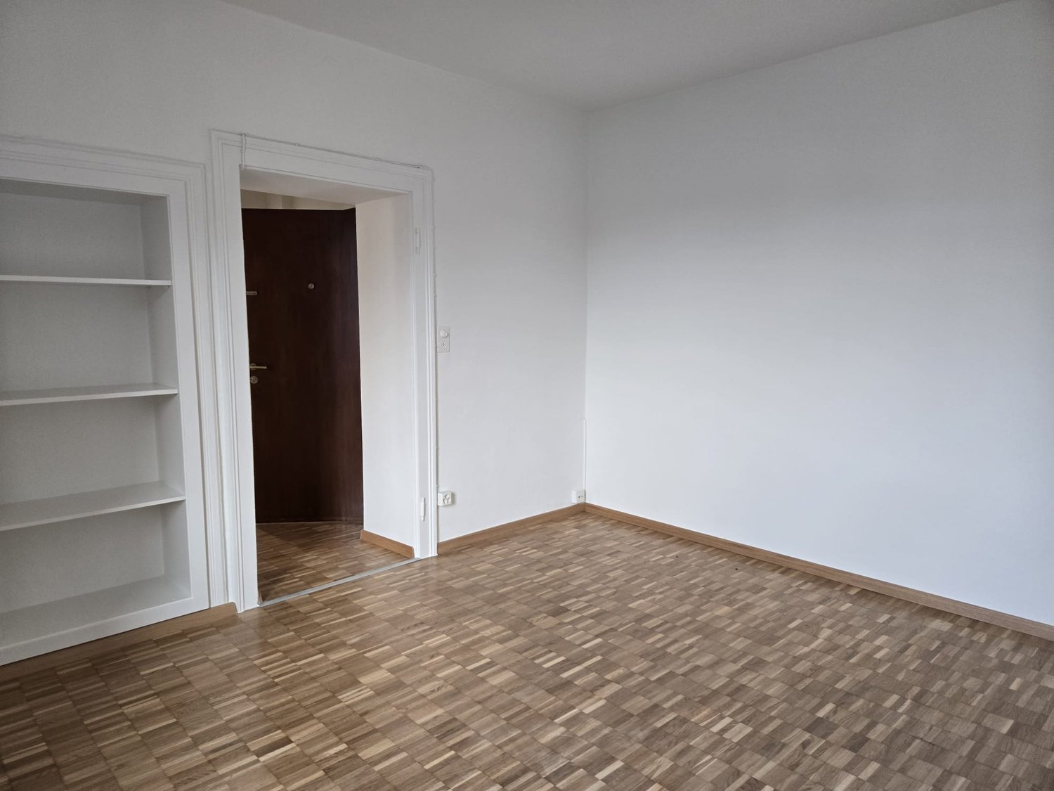 empty room with white walls, wooden parquet flooring, door, white built-in shelves