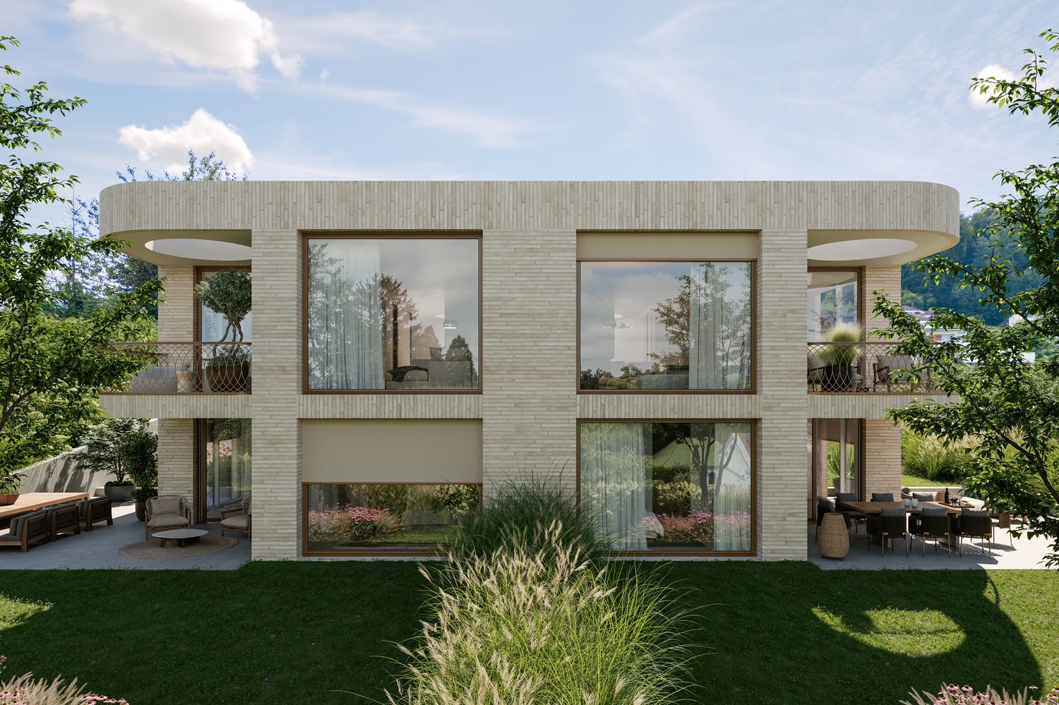 Two-story modern house with a flat roof, large windows, and a balcony or terrace. The exterior is made of light-colored bricks or stone. The house is surrounded by a well-landscaped garden with lush greenery.