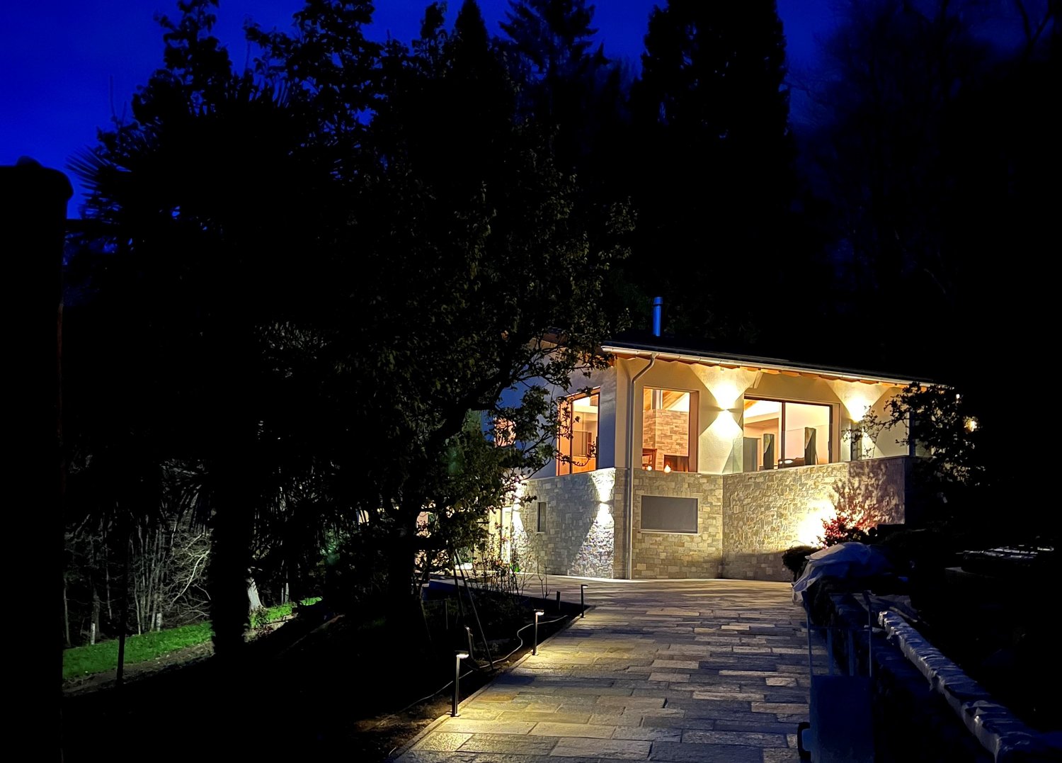 A modern house with a stone wall and glass windows, with lights on at night.