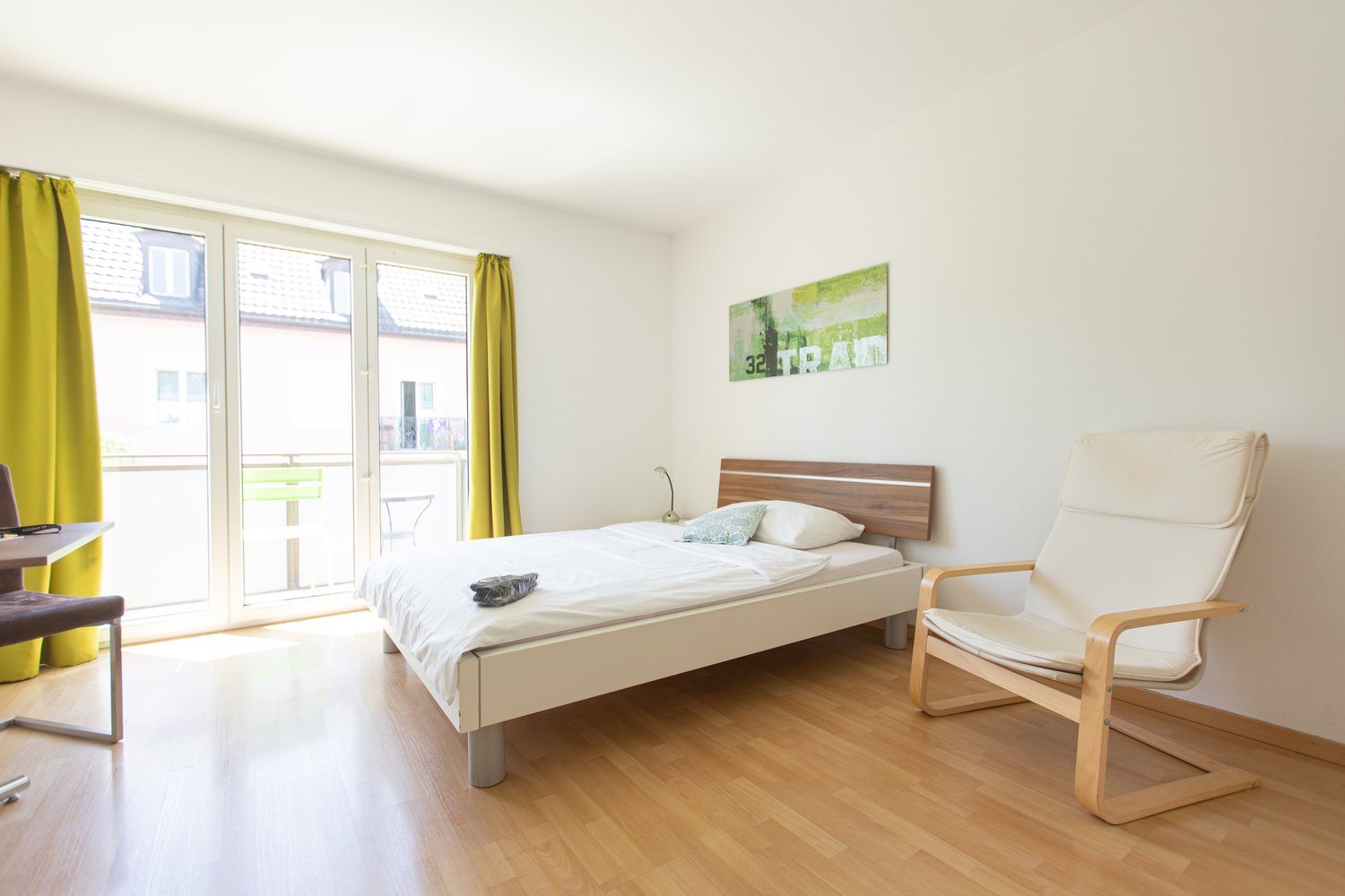 Spacious bedroom with large windows, wooden floor, white walls, a bed with wooden headboard, a comfortable armchair, and a colorful artwork on the wall.