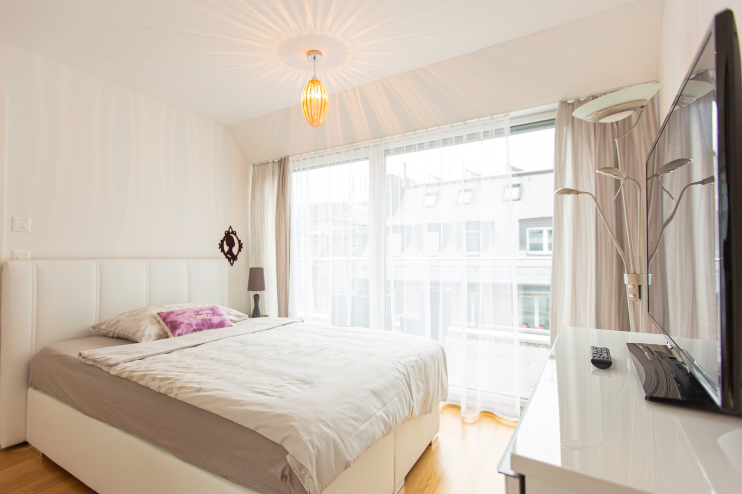 white bed, white bedhead, white pillow, brown blanket, lamp, tv, wooden floor