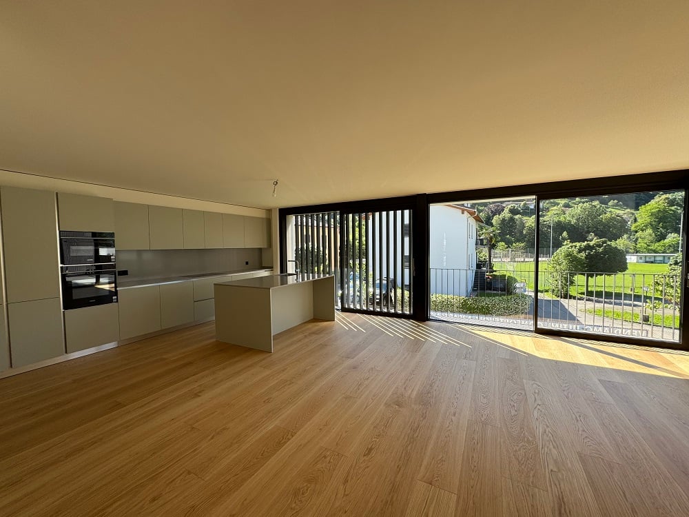 Spacious open-plan living area with hardwood flooring, modern kitchen with built-in appliances, and large sliding glass doors leading to a balcony or terrace overlooking a green outdoor area.