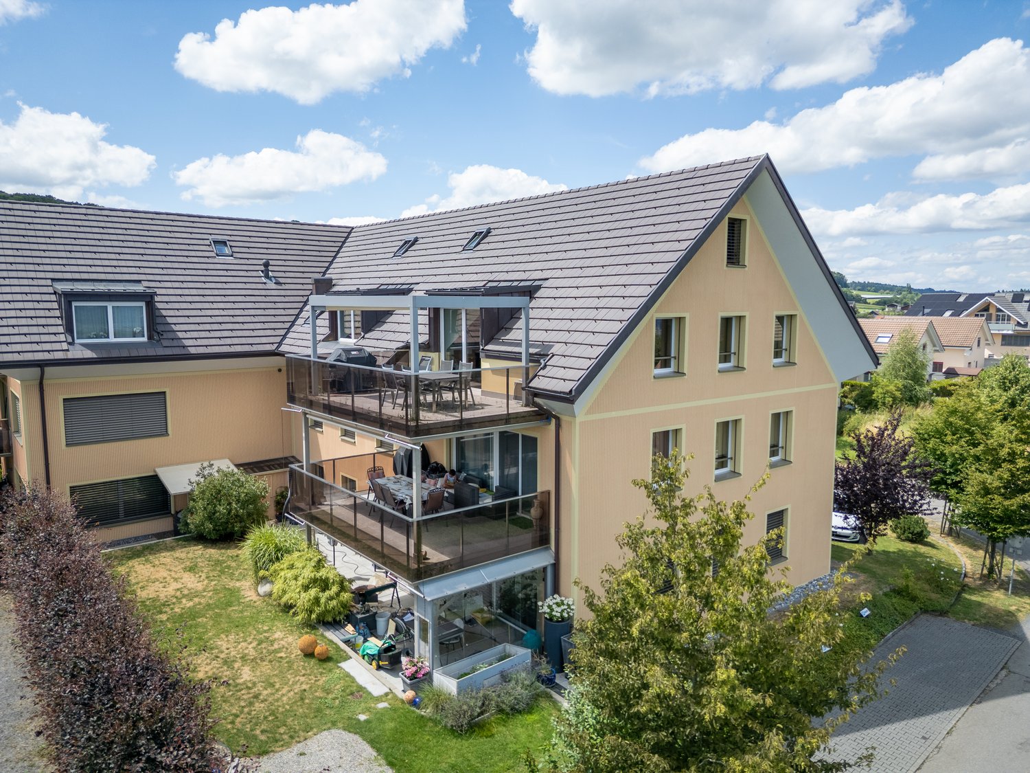 two story building, beige exterior, gray roof, balcony with glass railing, car parked on driveway, garden area with green grass and plants, trees surrounding the property