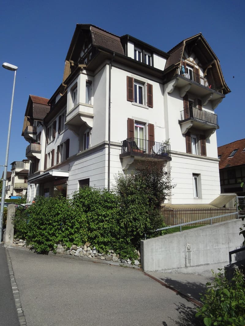 White building with brown roof, balconies, gables, multiple windows, shrubs in front