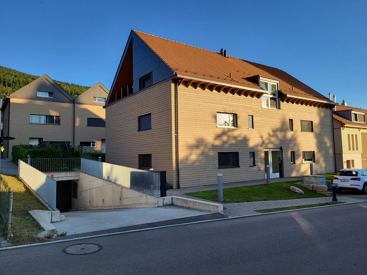 3-story apartment building with orange tiled roof, wooden siding, and large windows. There is a parking area in front of the building.