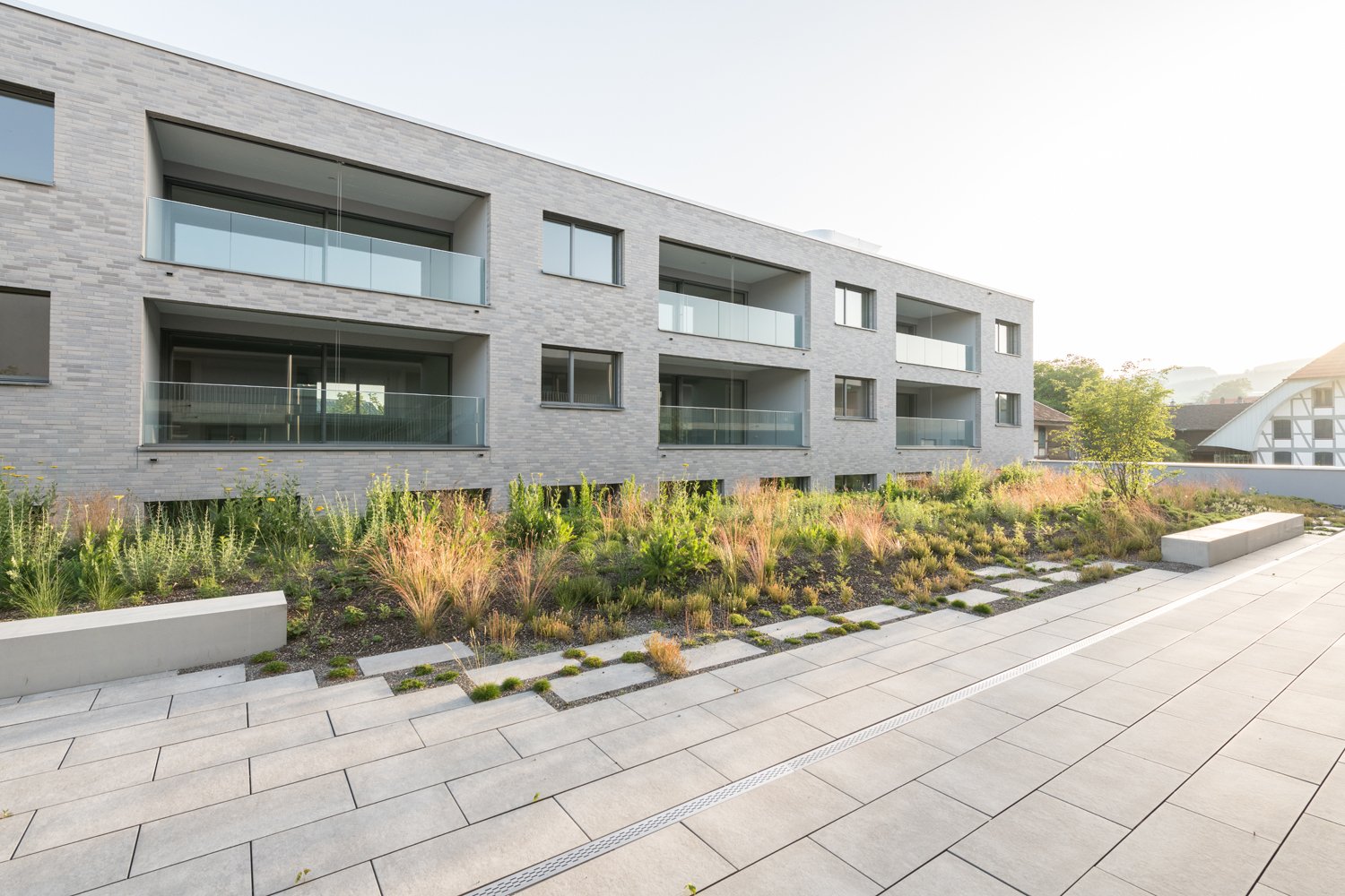 Multi-story apartment building with gray brick exterior, large windows, and balconies. The building is surrounded by landscaped gardens and walkways with stone pavers.