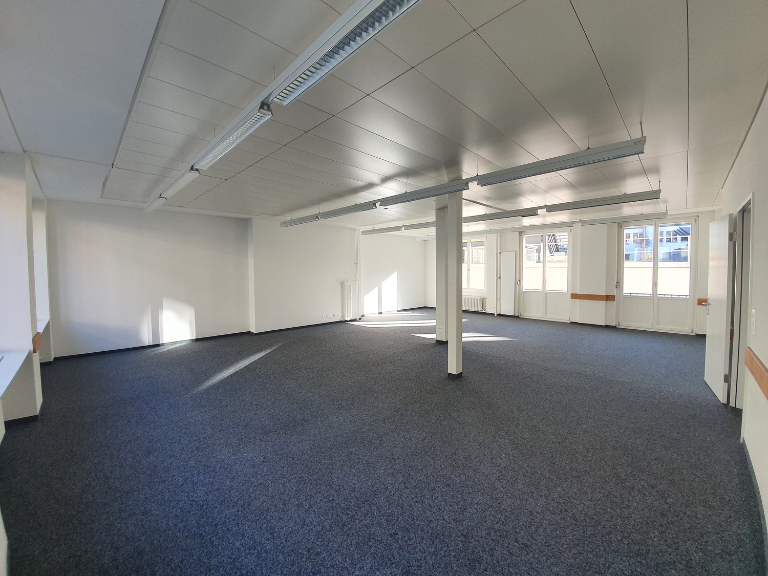Large open office space with high ceilings, white walls, and gray carpeted flooring. The room has several support columns and large windows providing natural light. The space appears to be ready for use as an office or commercial space.