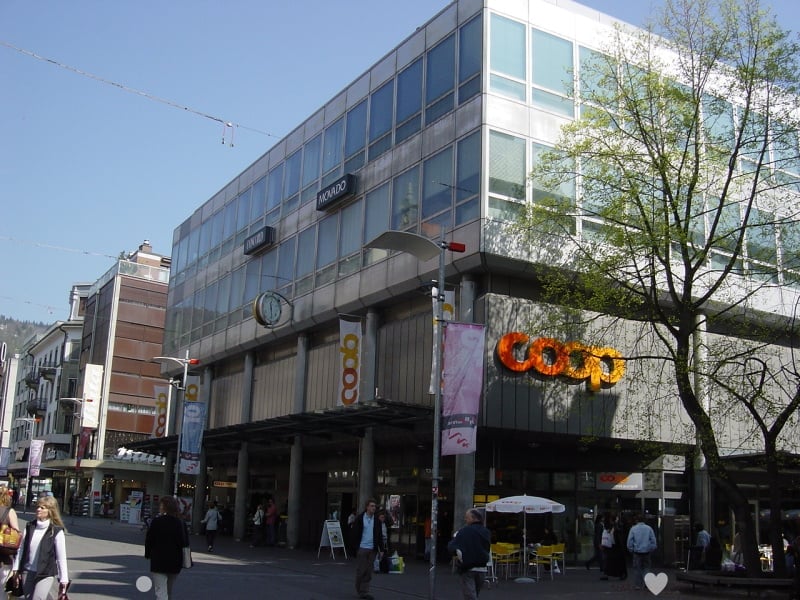 Modern building with glass facade, large sign reading 'COOP', multiple floors, people walking in front