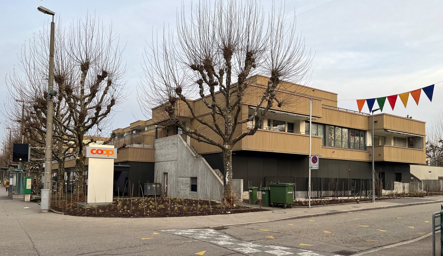 A modern three-story building with a gray-brown facade, large windows, and a sloped entrance. It includes a basement level with a ramp, a second floor balcony, and a flat rooftop with a small flag display.