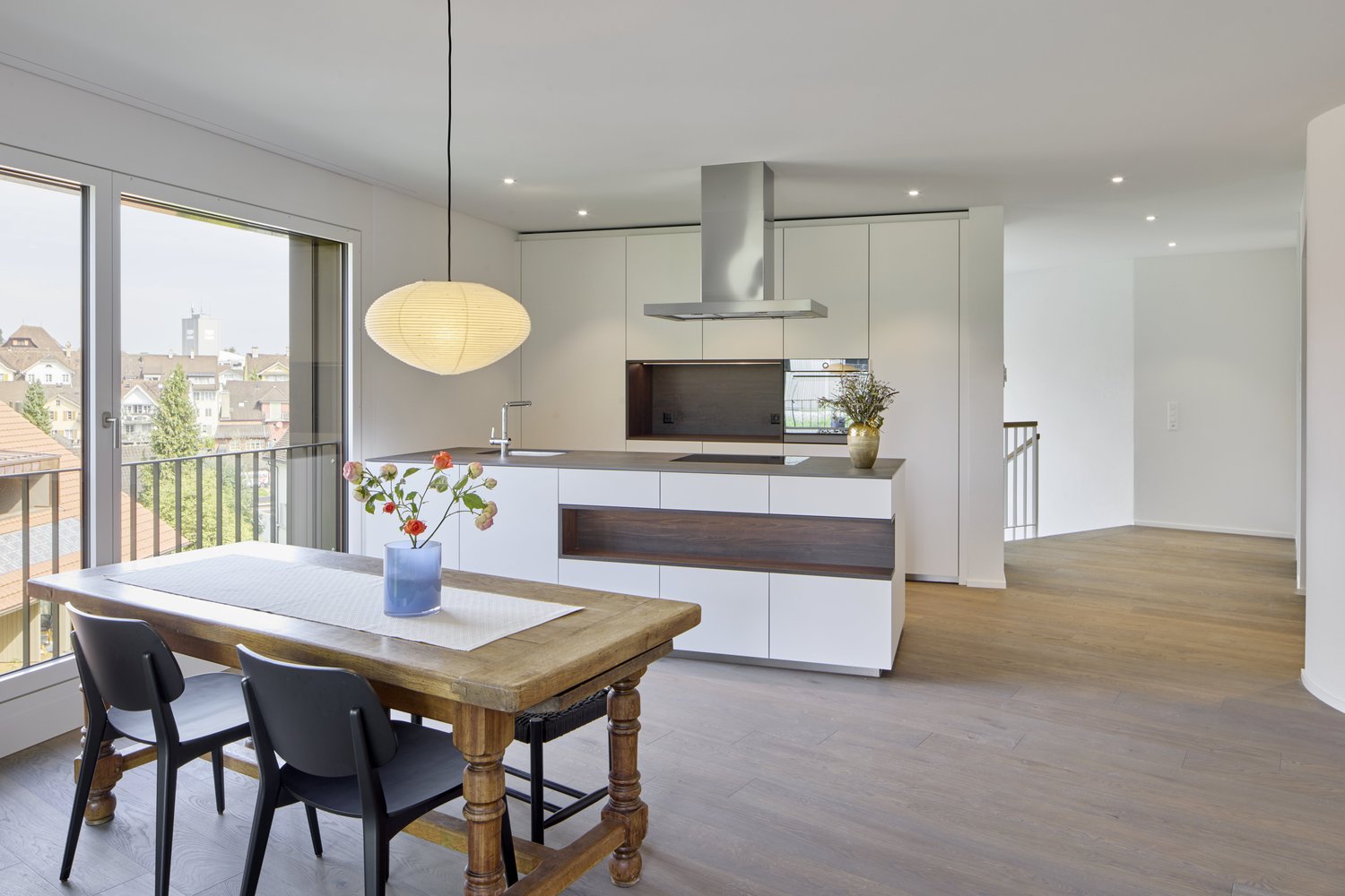 Modern kitchen with dining area, large windows, wooden floor, table and chairs, hanging lamp, stainless steel range hood, flowers in vase