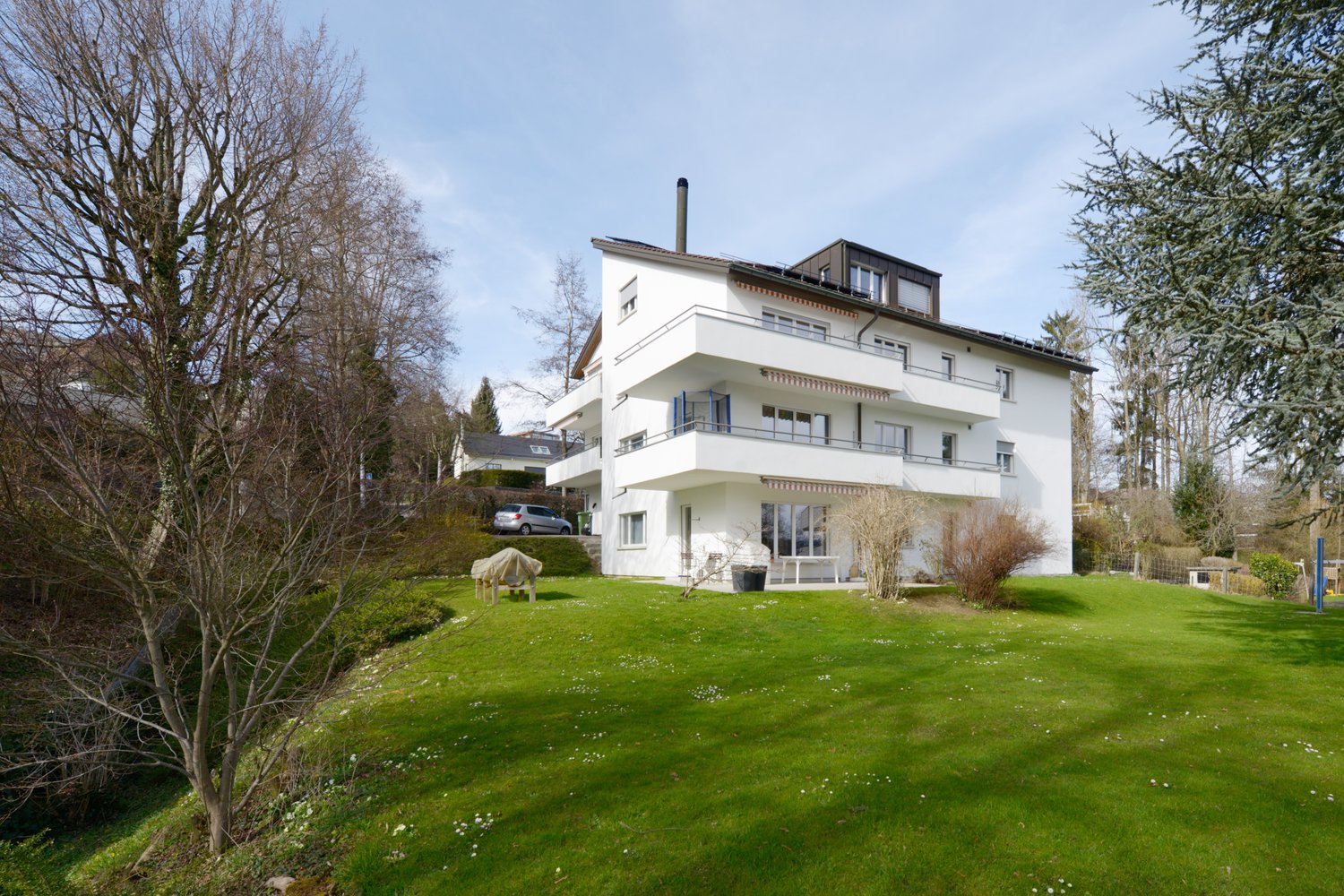 3-story white modern house with balconies, surrounded by a well-maintained lawn and trees, with a parking space visible in the front