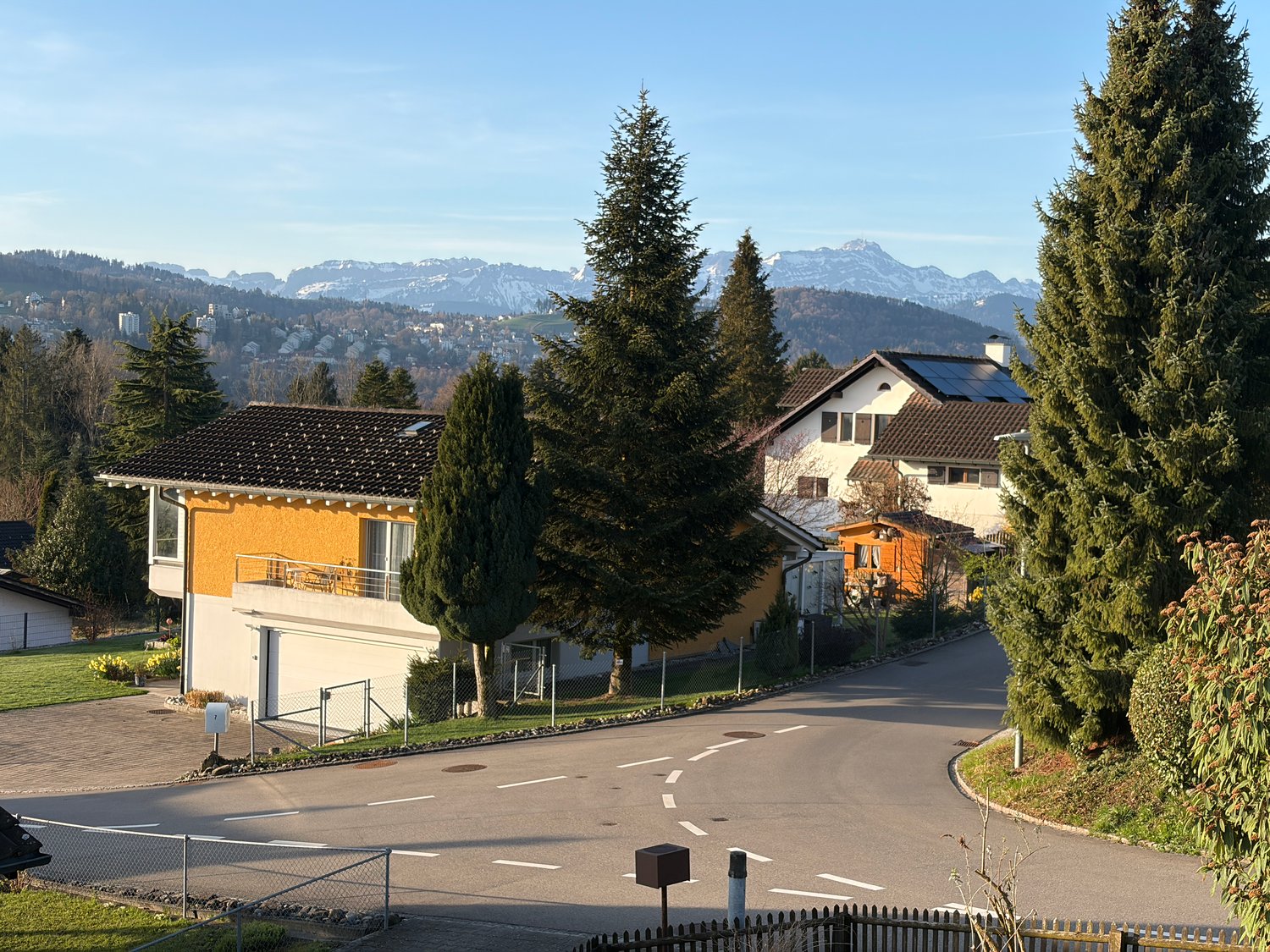 House with brown roof, yellow walls, balcony, solar panels, garage, driveway, trees, mountains in the background