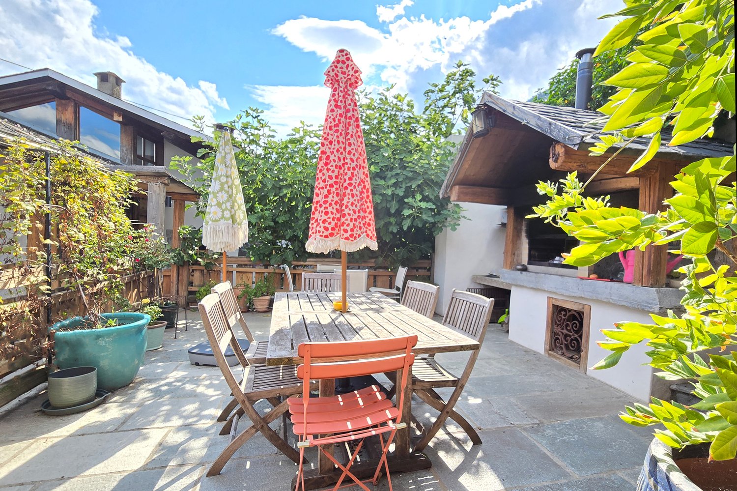 Outdoor dining table with chairs, 2 umbrellas, potted plants, and a wooden barbecue, clear sky