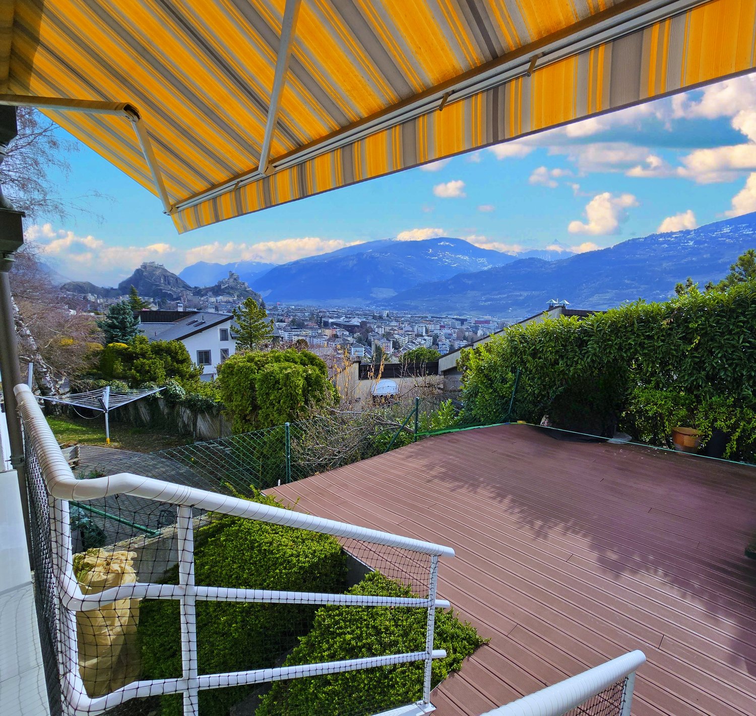outdoor terrace with awning, railing, wooden floor, bushes, net, cityscape view, mountains in the distance