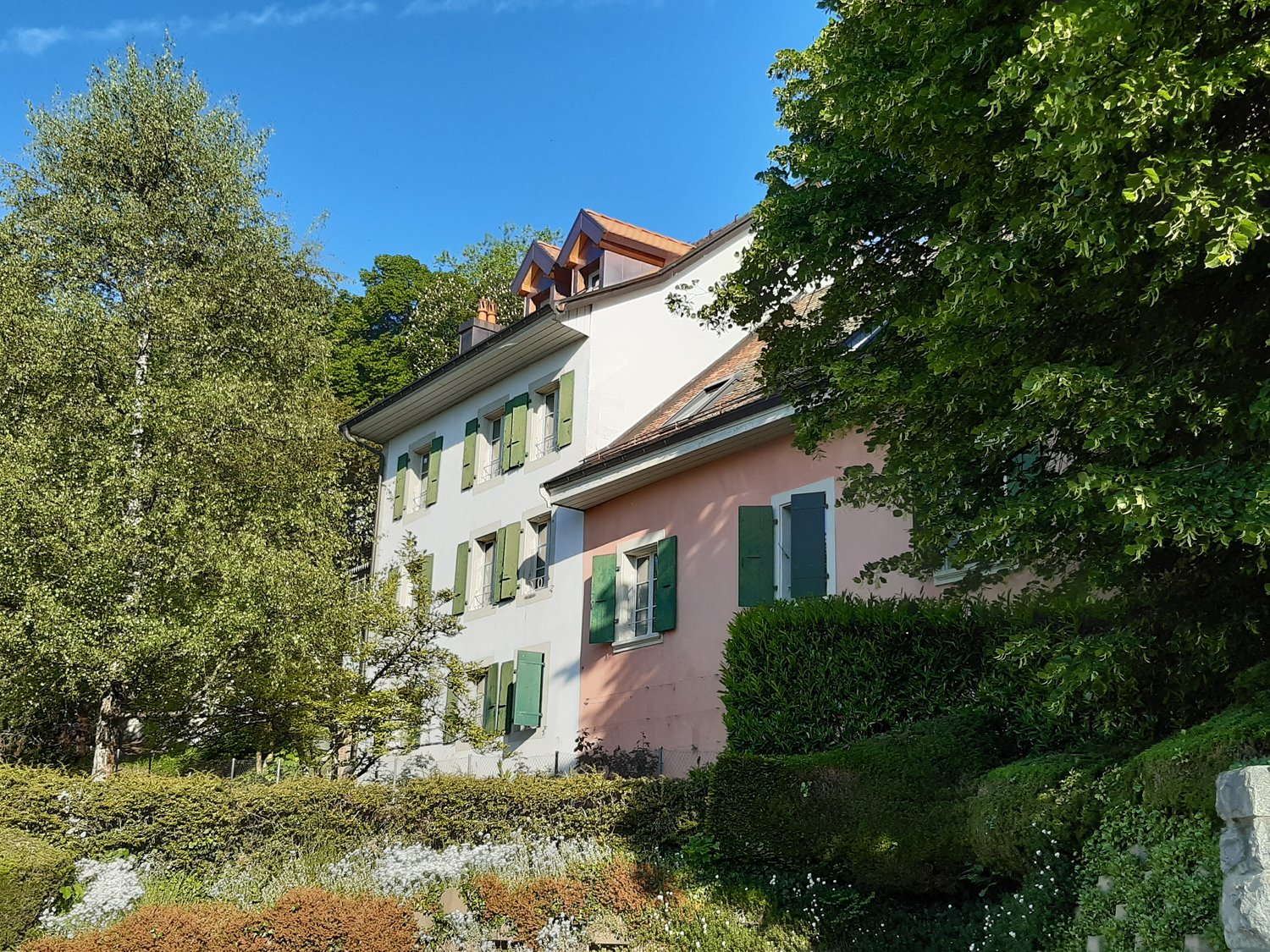 Two story house, white and pink facade, green shutters, red roof, surrounded by trees and bushes, garden area