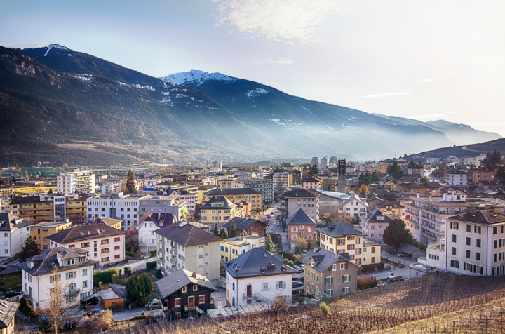 Panoramic view of a mountainous city with colorful buildings, surrounded by snow-capped peaks and hazy mountains in the background