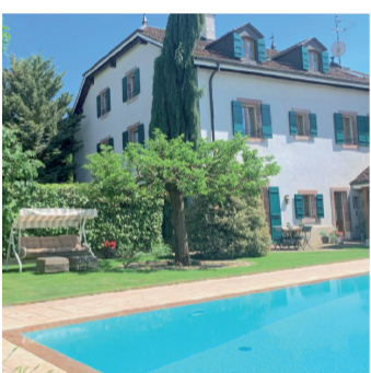 Two-story house with white exterior, red roof tiles, and green shutters. Large swimming pool in the foreground, surrounded by a paved area and lush greenery. There appears to be a balcony or terrace on the upper level of the house.