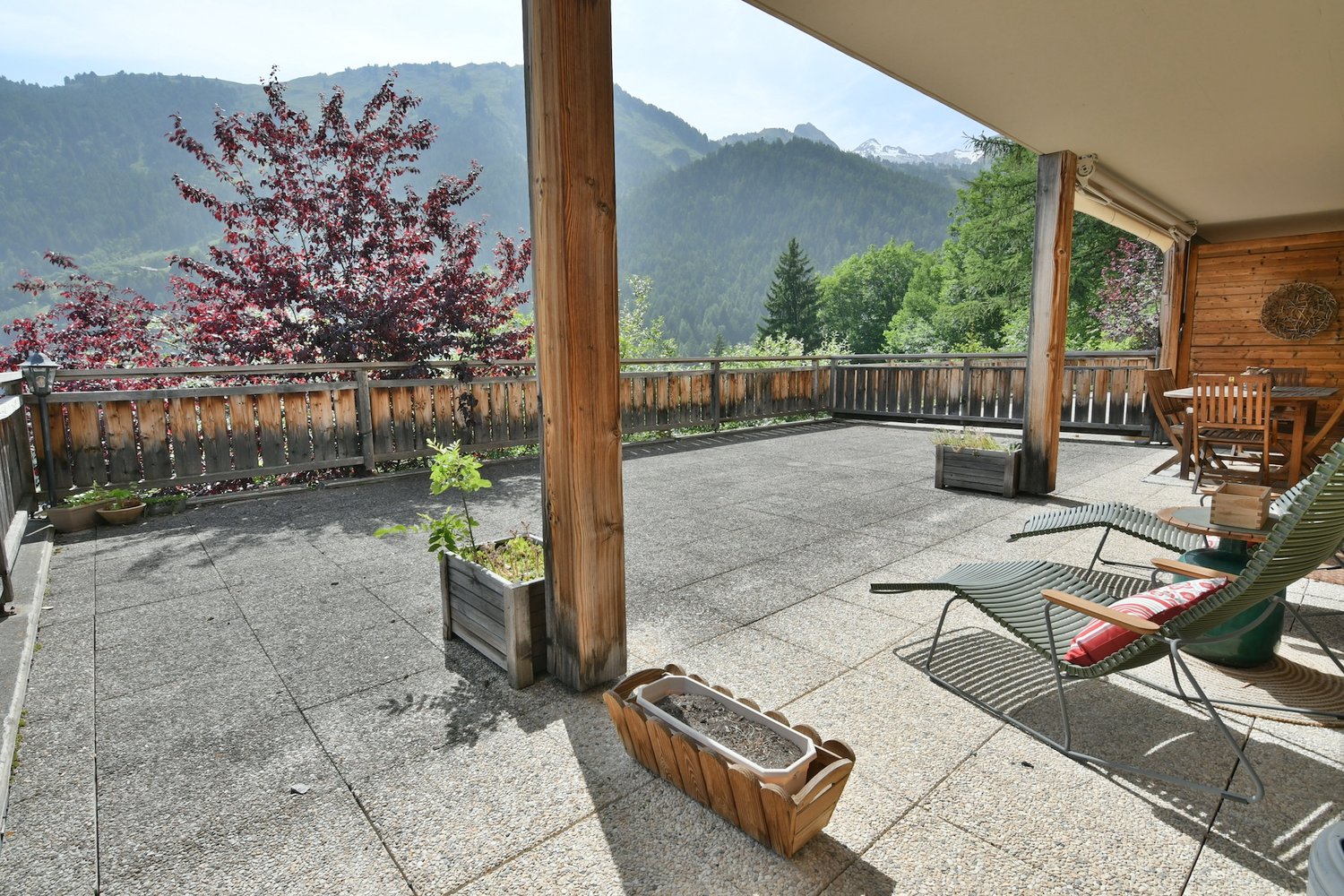 Wooden balcony or terrace with a scenic view of the mountains and forests. The balcony has a concrete floor, wooden railings, and some potted plants. There are chairs and other outdoor furniture on the balcony, suggesting it is a relaxing outdoor living s