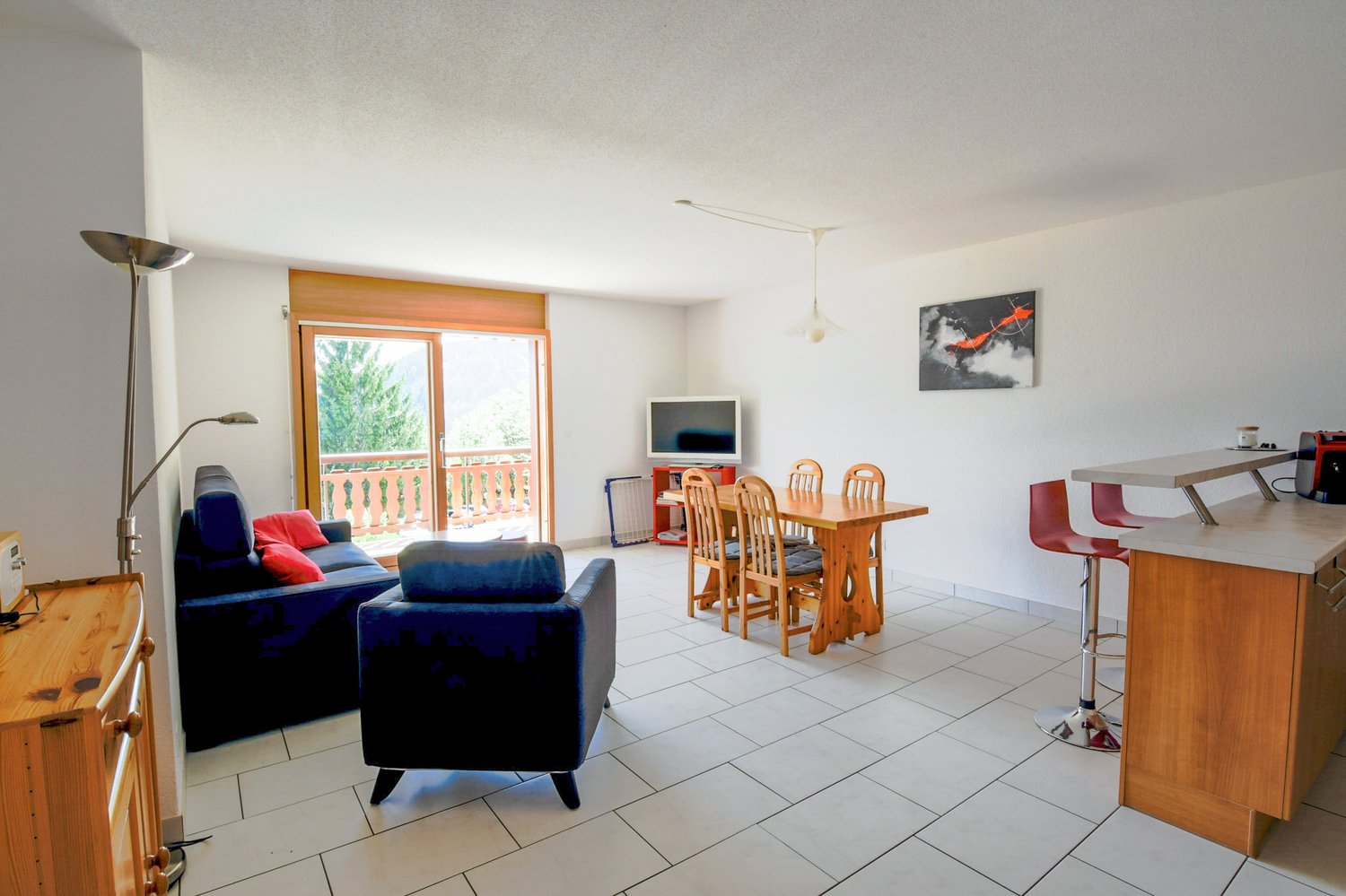 Open-plan living room and dining area, with large windows leading to a balcony or terrace. The room has tiled flooring, a wooden dining table with chairs, and a sofa and armchair in a blue color scheme. There is also a TV and other electronic equipment vi