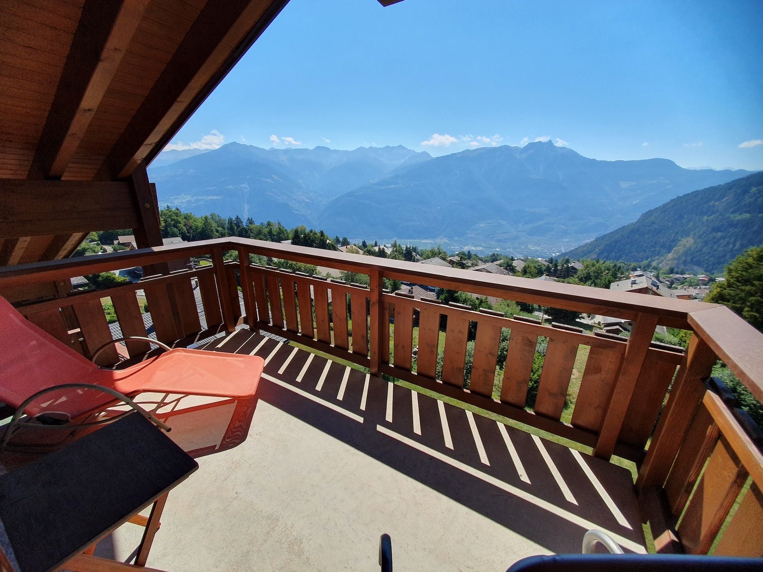 wooden balcony with lounge chair, wooden railing, panoramic mountain view, grassy slope below