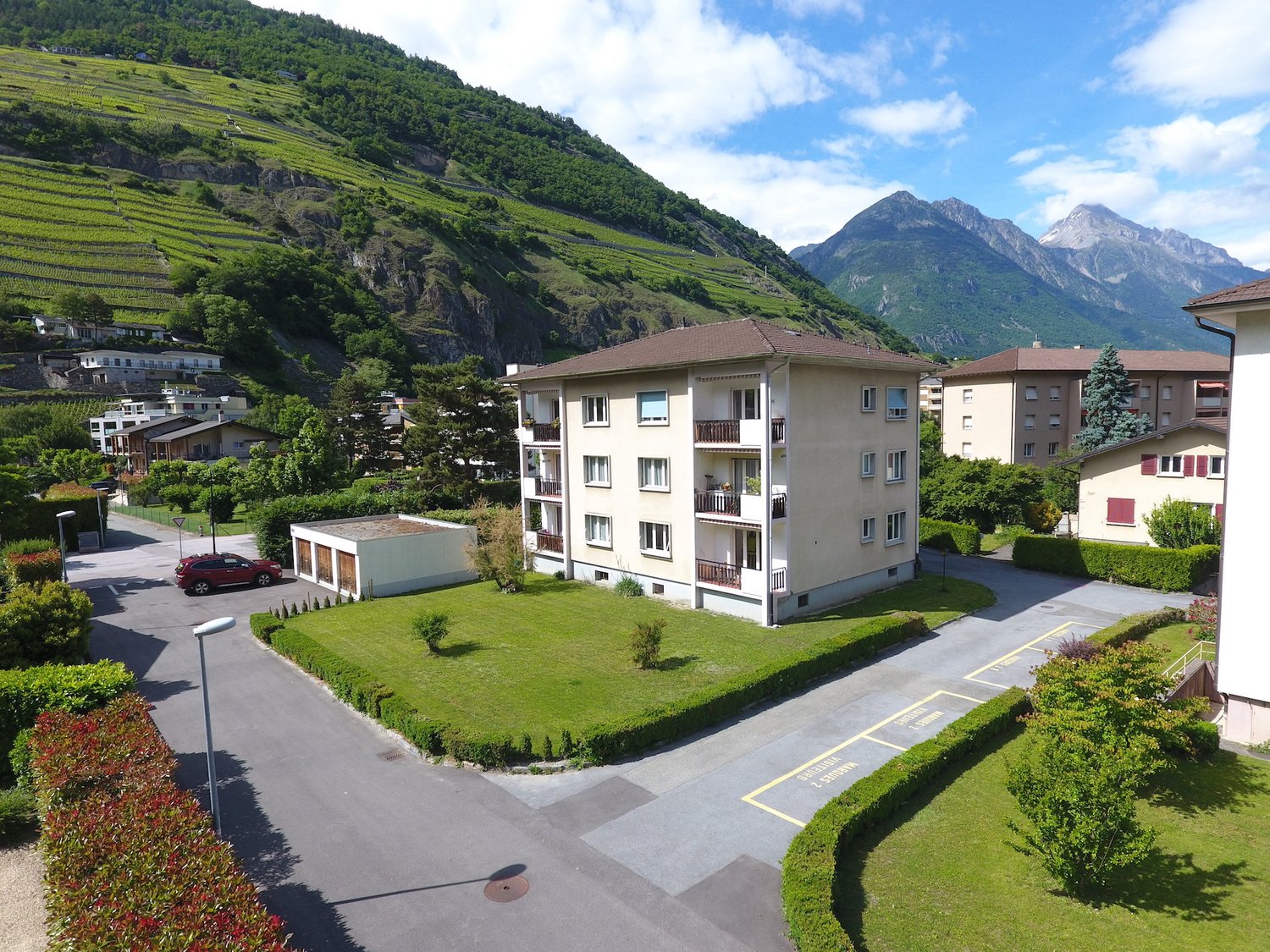A multi-story apartment building with balconies, surrounded by well-maintained landscaping and parking spaces. The building is situated in a scenic mountainous area with lush green hills and peaks in the background.