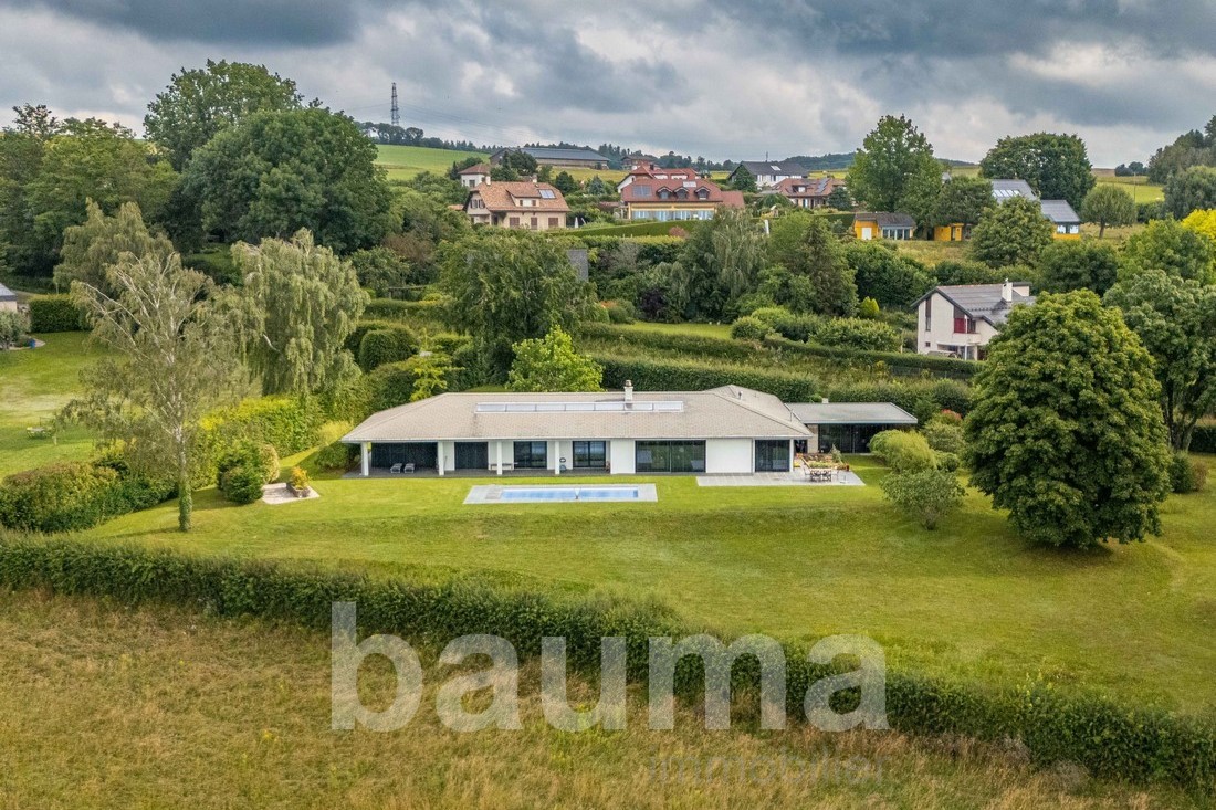 Single-story modern house with a swimming pool, surrounded by lush greenery and trees. The house is situated in a scenic rural area with other residential buildings visible in the background.